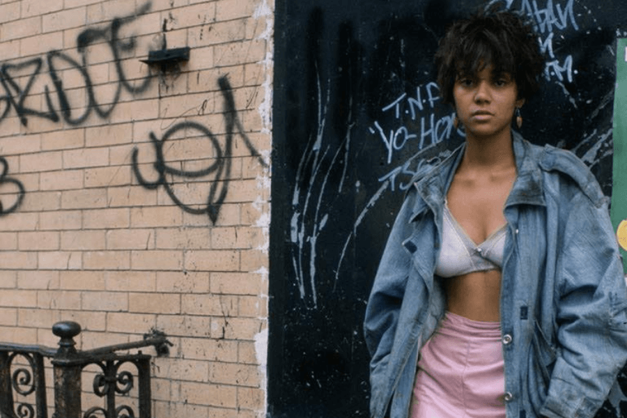 This image shows a young woman standing against a graffiti-covered wall, wearing a distressed denim jacket over a bra and a light pink skirt. Her expression appears serious, and the urban setting conveys a gritty and raw atmosphere.