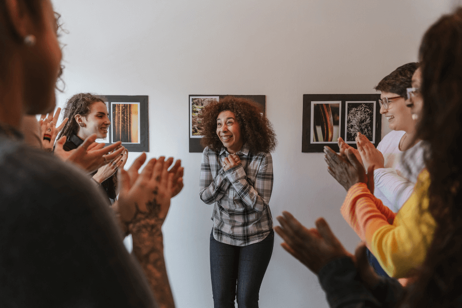 A woman stands smiling in the center of a group of people clapping and celebrating her. The setting appears to be indoors, with framed artwork on the wall in the background.