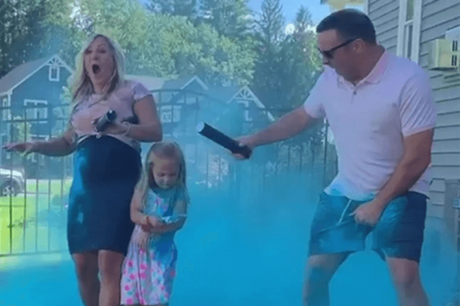 This image shows a joyful gender reveal celebration where a couple and a young child are surrounded by a cloud of blue powder, indicating a baby boy. The woman appears surprised and excited, while the man is enthusiastically holding a confetti cannon. The scene is set in a backyard with a fence and houses visible in the background.