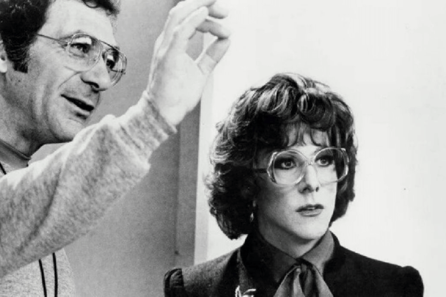 This black-and-white image shows a man speaking and gesturing beside a person wearing large glasses, a wig, and a blouse with a bow tie. The person in the wig has an attentive, serious expression, suggesting a behind-the-scenes or on-set moment.