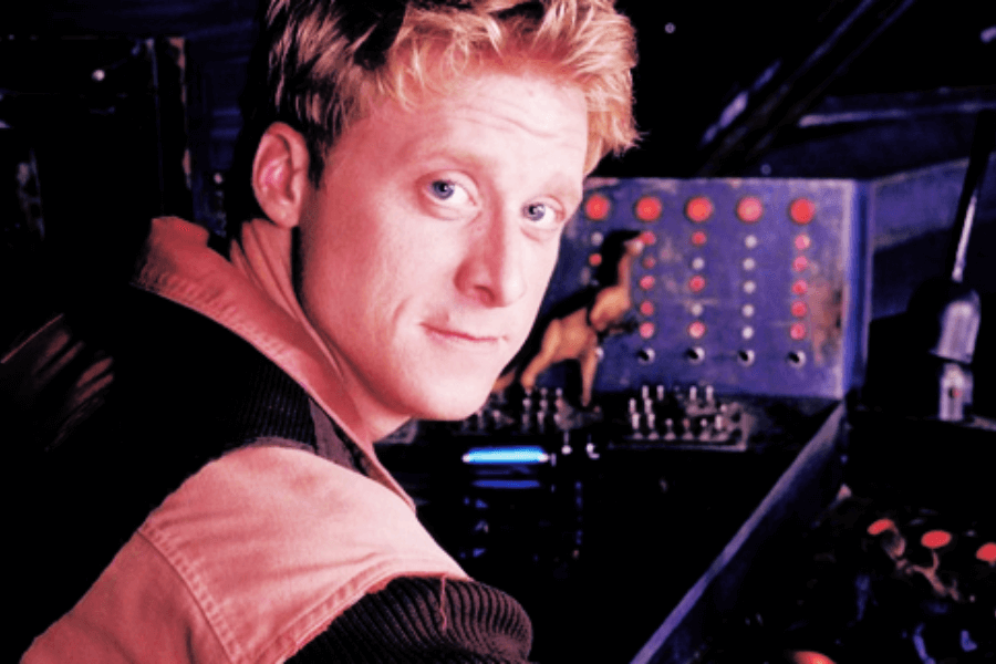 This image features a man with short blonde hair and a friendly, relaxed expression, seated at a control panel with various buttons and switches. A small toy dinosaur is visible on the panel, adding a playful touch to the futuristic or mechanical setting. The lighting and focus suggest a casual yet intriguing moment in a high-tech environment.