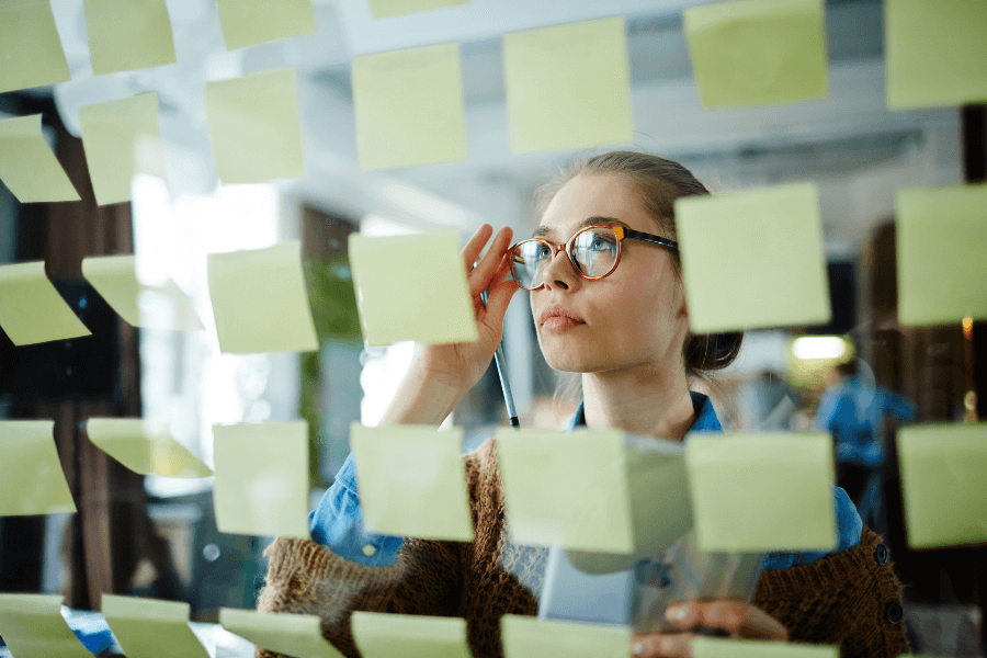 A woman wearing glasses thoughtfully examines a wall or glass panel covered in yellow sticky notes. She holds a pen and a sheet of paper, appearing focused in a well-lit workspace.