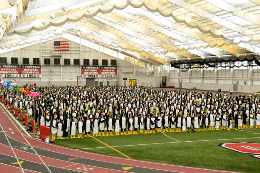 The image shows a large indoor gathering of people dressed in penguin costumes, standing in organized rows on an athletic field. The venue is a spacious indoor sports facility, with a track surrounding the field and banners displaying football championship years on the walls. This appears to be a coordinated event, possibly aiming for a Guinness World Record or similar group achievement involving the most participants in penguin attire.