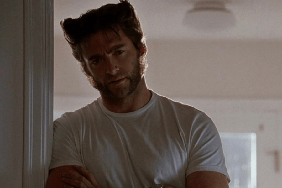 This image shows a man with distinctive, spiked hair and prominent sideburns, standing casually in a doorway with his arms crossed. He is wearing a plain white T-shirt and has a thoughtful, slightly stern expression. The softly lit, neutral-toned background adds a calm yet introspective mood to the scene.