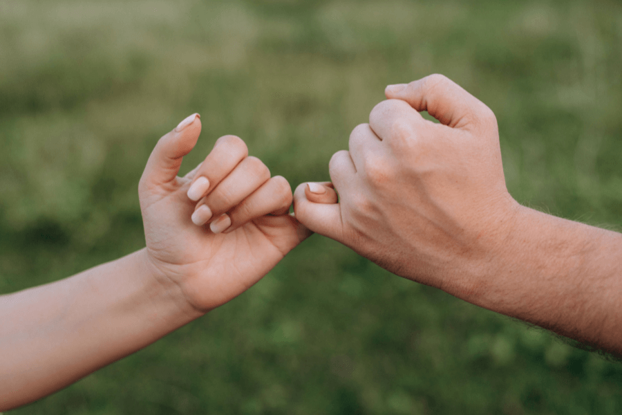 Two hands linking pinky fingers against a blurred green outdoor background, symbolizing a promise or bond.