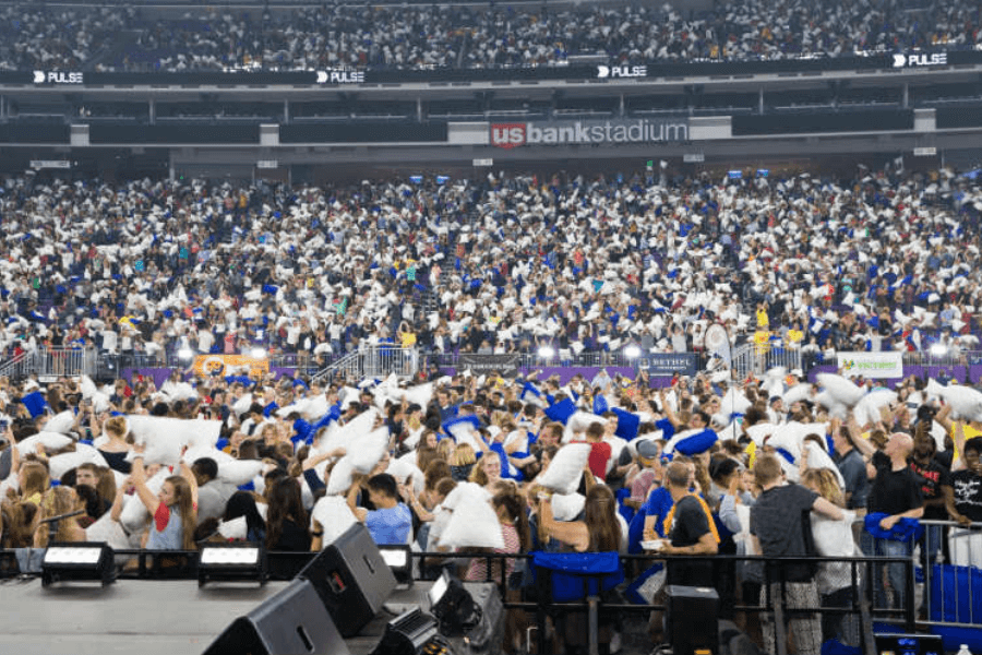 The image shows a massive gathering of people participating in a large-scale pillow fight at the U.S. Bank Stadium. The crowd is packed with participants waving and swinging pillows, creating a festive and chaotic scene. The stadium's branding and a lively atmosphere suggest this event is a coordinated attempt to set a Guinness World Record for the largest pillow fight or a similar achievement.