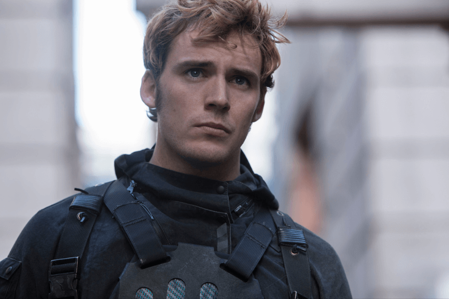 This image features a young man with blonde tousled hair, wearing a dark tactical outfit with straps and armor, standing in an urban setting. His serious expression suggests focus or concern, and the blurred buildings in the background create a sense of action or preparation. The lighting highlights his determined demeanor.