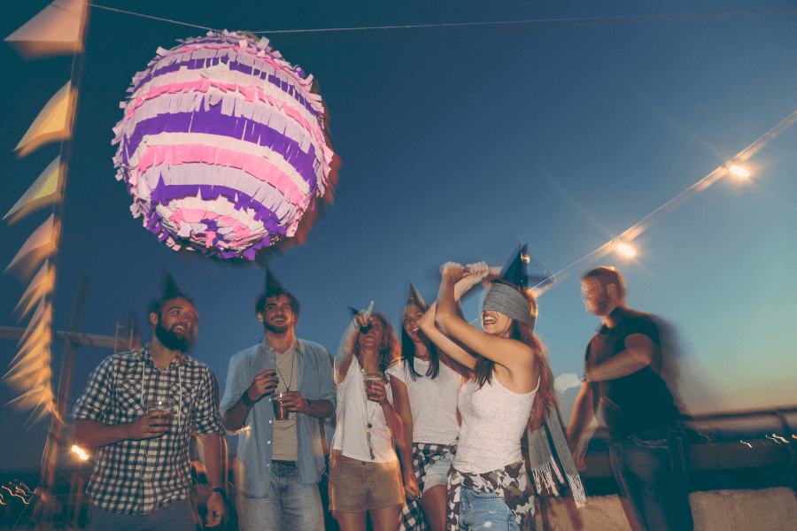 This image captures a festive outdoor party at sunset, with a group of people gathered under string lights. A blindfolded woman swings a stick at a colorful piñata while others watch and cheer, wearing party hats. The joyful atmosphere is complemented by warm lighting and vibrant decorations.