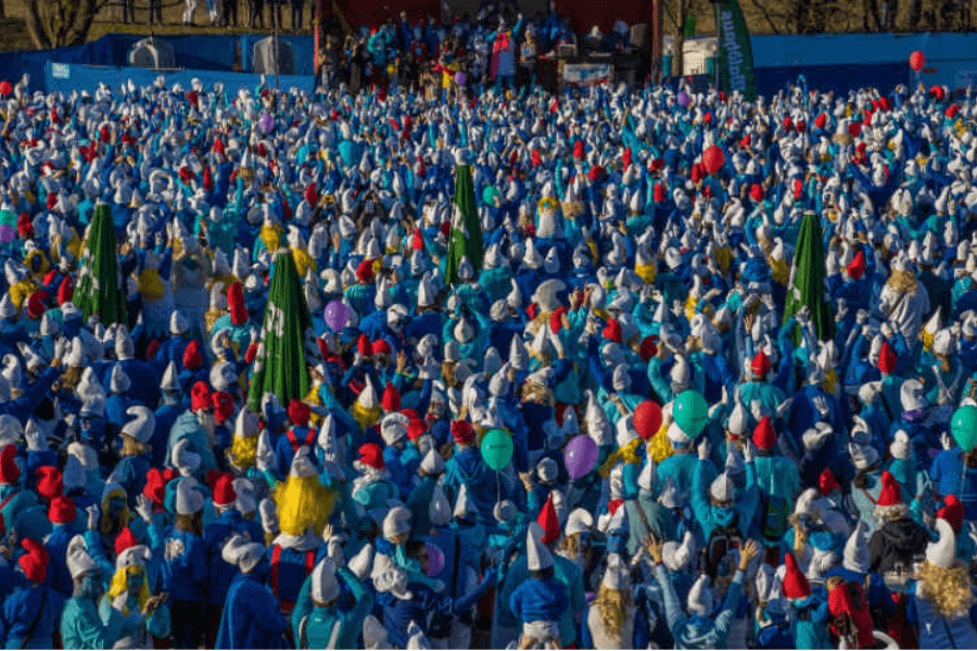 The image captures a vibrant outdoor gathering of thousands of people dressed as Smurfs, wearing blue outfits and white or red hats. Balloons and festive decorations are visible among the crowd, adding to the celebratory atmosphere. This appears to be a large-scale event, likely an attempt to set or break a Guinness World Record for the largest assembly of people dressed as Smurfs.