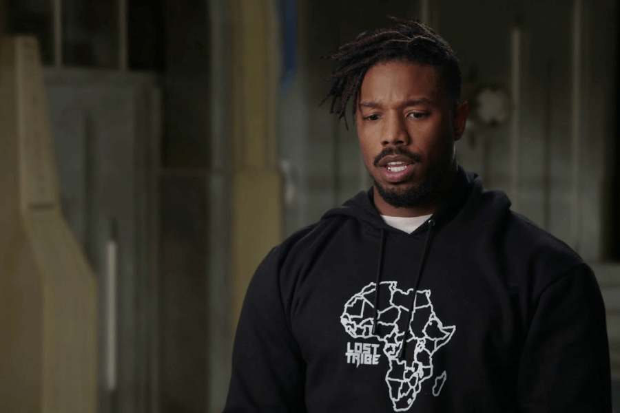 This image shows a man wearing a black hoodie with a graphic of the African continent and the text "Lost Tribe" printed on it. He has a thoughtful expression and stands in an indoor setting with muted, industrial-style surroundings.