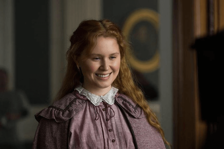 This image features a young woman with long, wavy red hair, smiling warmly while wearing a period-style outfit with a lace collar and a pinkish-purple dress. The setting appears to be indoors, with blurred background elements such as a framed portrait and columns, suggesting a historical or elegant atmosphere. Her expression conveys warmth and charm, enhancing the overall serene tone.