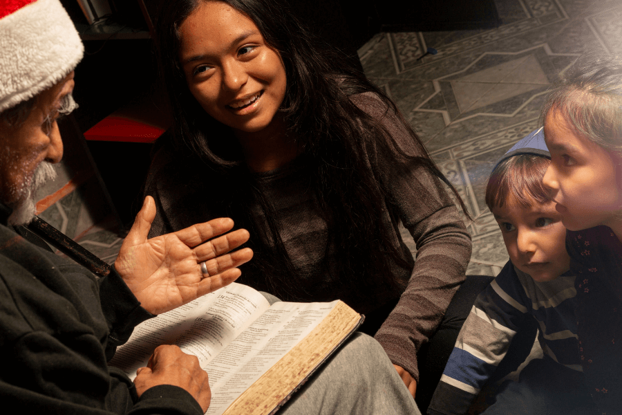 An elderly man wearing a Santa hat reads from an open book to a woman and two children who listen intently. They are gathered on a tiled floor, creating a warm and intimate storytelling moment.