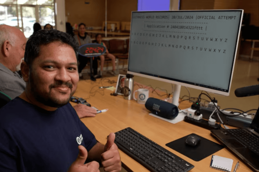 The image shows a man smiling and giving a thumbs-up during a Guinness World Records attempt. He is seated at a desk with a keyboard, and a computer screen in front of him displays text related to the official record attempt, dated 20 July 2024. In the background, other participants and onlookers are visible, creating a supportive and formal atmosphere for this record-setting activity.