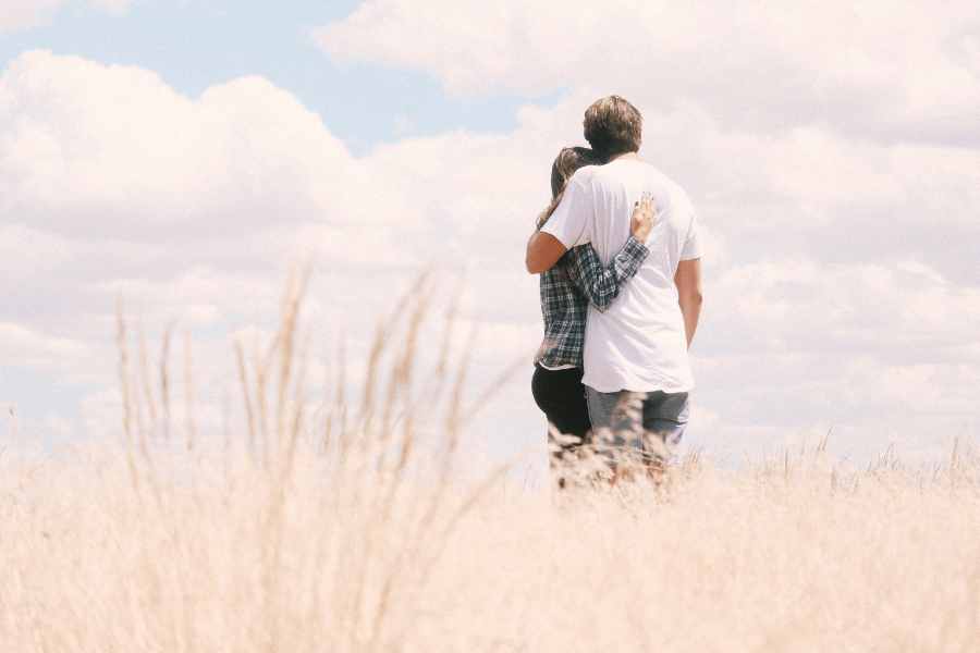 A couple stands in a field of tall grass, embracing as they look into the distance under a bright blue sky with scattered clouds.