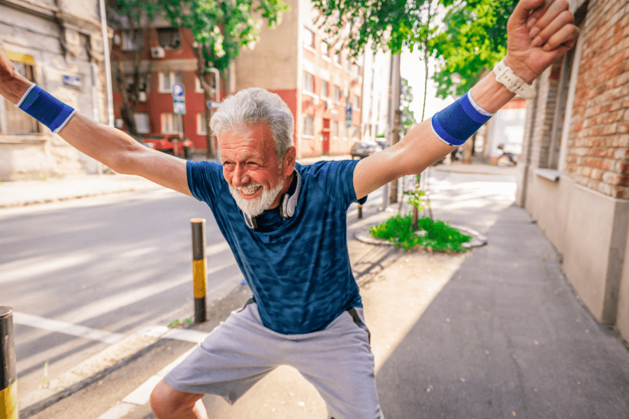 An energetic elderly man with a big smile stretches his arms in celebration while standing on a city sidewalk. He is dressed in athletic wear with sweatbands and headphones, exuding joy and vitality.