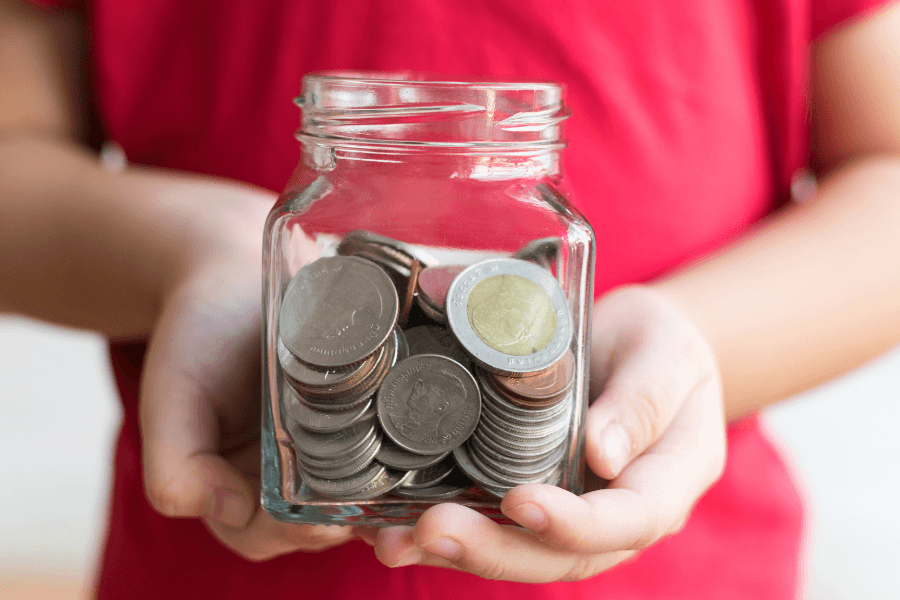 A person holding a clear glass jar filled with coins, including various silver and gold-toned pieces, with a red shirt visible in the background.