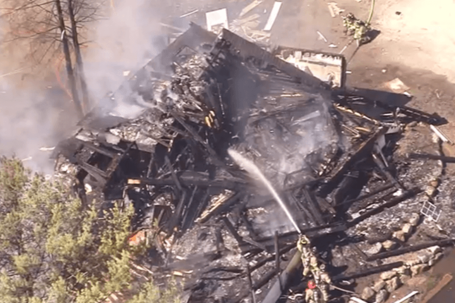 This image shows the aftermath of a large fire, with firefighters spraying water on the smoldering remains of a collapsed structure. The area is surrounded by charred debris and scorched earth, with smoke still rising from the scene.