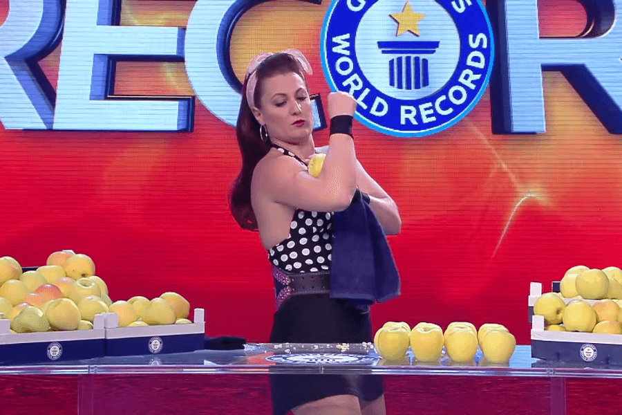 The image shows a woman in a polka dot top and vintage-style headband participating in a Guinness World Records event. She is polishing a yellow apple against her arm with a cloth, standing behind a table covered with neatly arranged apples and boxes filled with more apples. The backdrop prominently features the Guinness World Records logo and the word "RECORDS" in bold letters.