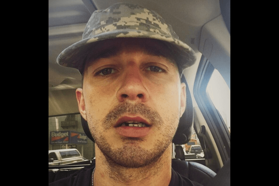 This image shows a close-up of a man sitting in a car, wearing a digital camouflage-patterned cap. He has a slight stubble and a neutral expression, with the background revealing parts of the car interior and a parking lot outside.