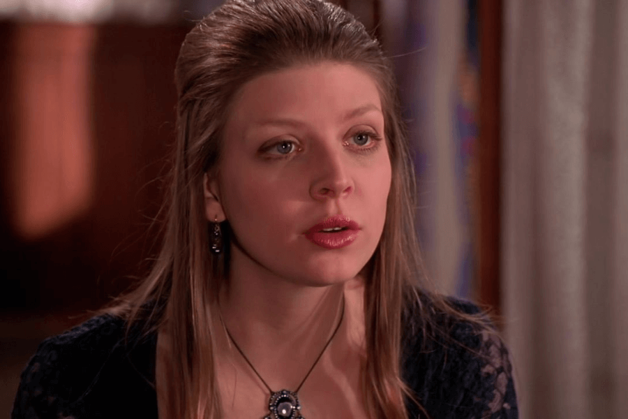This image shows a young woman with long, light brown hair, styled back from her face, wearing elegant earrings and a pendant necklace. She has a thoughtful and slightly emotional expression, suggesting a heartfelt or serious moment. The warm indoor background with soft lighting adds a sense of intimacy and focus to her demeanor.