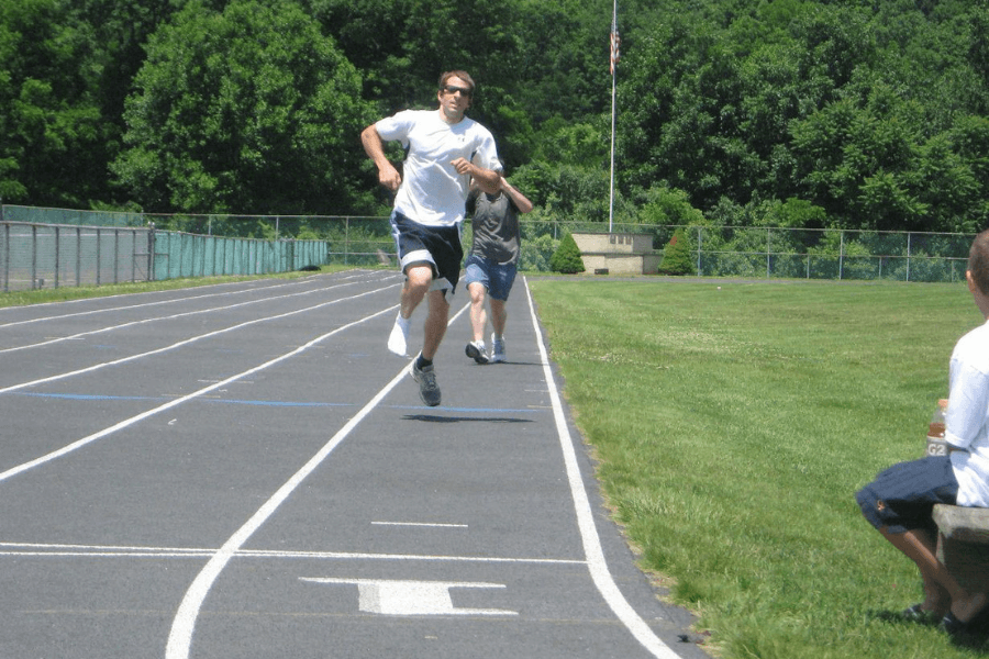 The image shows a man running at high speed on an outdoor track, wearing a white shirt and dark athletic shorts. He appears to be competing or training, with another person running behind him and spectators observing from the side. The setting is a sunny day, surrounded by green trees and grass, creating a casual yet competitive atmosphere. The focus is on the lead runner’s determined stride.