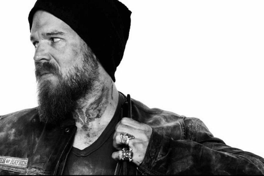 This black-and-white image features a rugged man with a thick beard and a tattooed neck, wearing a beanie and a leather jacket. He gazes to the side with a stern expression, gripping the lapel of his jacket. The monochromatic setting emphasizes his intense, edgy persona.