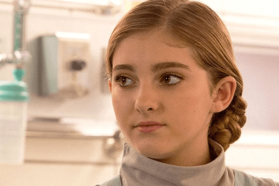 This image features a young woman with neatly braided blonde hair, wearing a high-necked outfit and gazing to the side with a calm, composed expression. The background includes blurred medical equipment, suggesting a clinical or scientific setting. Her focused demeanor conveys intelligence and attentiveness.