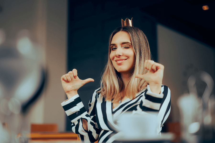 A woman wearing a striped outfit and a small crown gestures proudly with her thumbs pointing toward herself while winking. She exudes confidence and playfulness in a well-lit indoor setting.