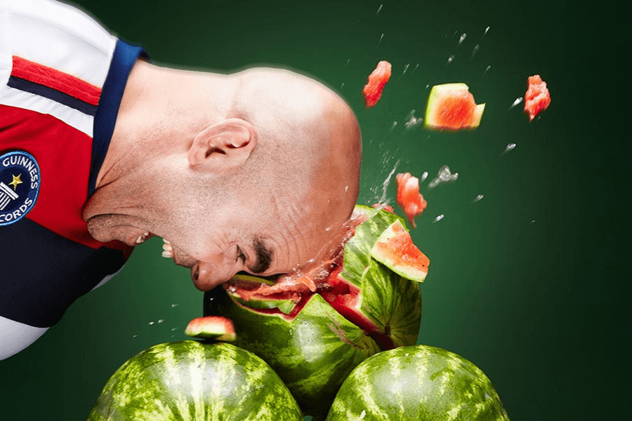 The image shows a man in action, smashing a watermelon with his head, captured mid-strike. Pieces of watermelon and juice explode outward, emphasizing the force of the impact. The man is wearing a shirt with a Guinness World Records logo, suggesting this is part of an official attempt or record-breaking event. The vivid green background and flying fruit pieces make the moment dynamic and intense.