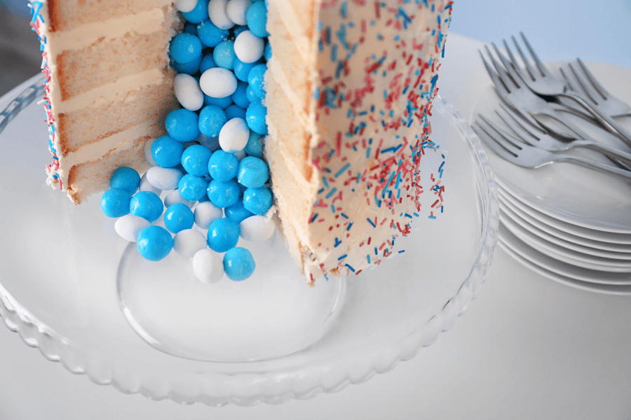 This image features a multi-layered cake covered in colorful sprinkles with a section cut out, revealing a cascade of blue and white candies spilling out from the center. The cake is displayed on a clear glass stand, with a stack of plates and forks neatly arranged nearby, suggesting a festive or celebratory occasion.