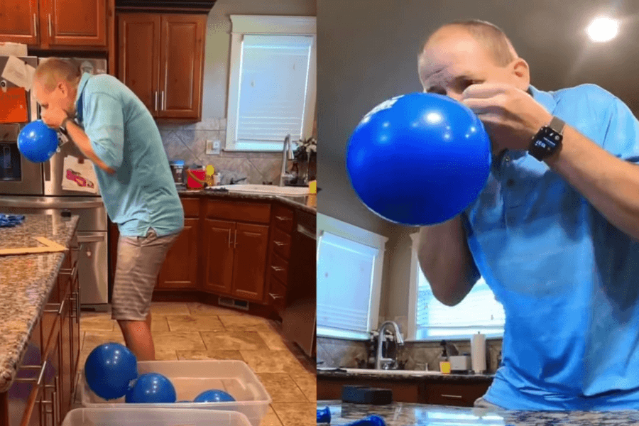 The image shows a split view of a man in a kitchen participating in a balloon-blowing challenge. On the left, he is standing and inflating a blue balloon, with several already-inflated balloons filling a plastic container at his feet. On the right, a close-up captures him intensely focused as he blows up another balloon. The casual home setting contrasts with the high-energy effort, suggesting this is part of a personal or record-breaking attempt.