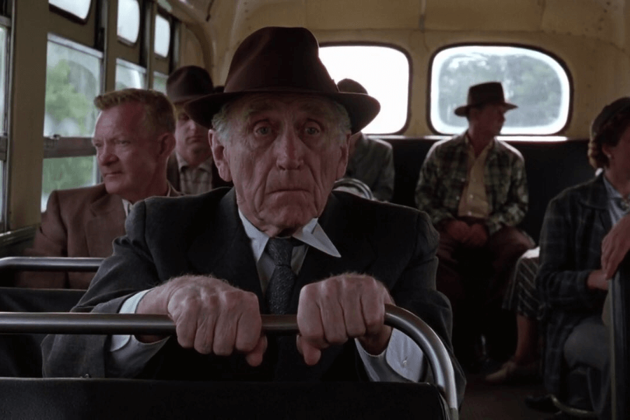 This image features an elderly man sitting on a bus, gripping the seat in front of him with both hands, wearing a dark suit and a brown fedora. His expression is solemn and introspective, while other passengers, dressed in mid-20th-century attire, sit in the background. The scene conveys a nostalgic or reflective mood, set in a simple and realistic environment.