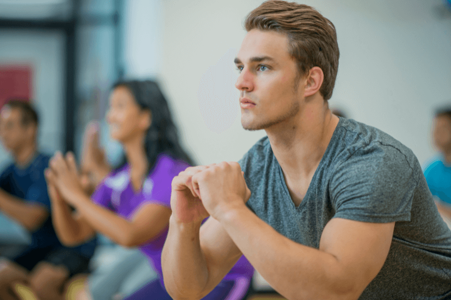 A man in a gray T-shirt focuses intently while participating in a group fitness class. Other participants, slightly blurred in the background, perform similar exercises in a brightly lit indoor setting.