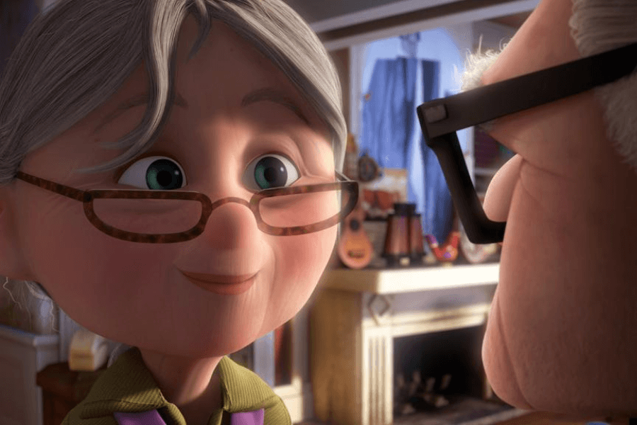 This image shows a close-up of an animated elderly woman with gray hair, round glasses, and a gentle smile, looking affectionately at someone nearby. The scene takes place in a cozy room with a fireplace, musical instruments, and warm lighting in the background, creating a heartfelt and intimate atmosphere. Her expression conveys love and tenderness.