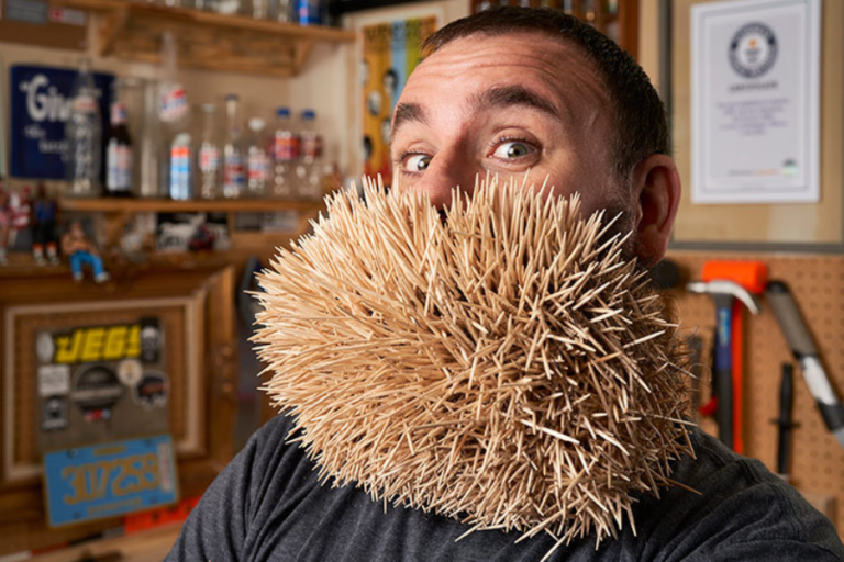 The image features a man posing with an extraordinary "beard" made entirely of toothpicks, creating a striking and humorous visual. His wide-eyed expression adds to the playful nature of the scene. The background shows a workshop or hobby room, with tools and a Guinness World Records certificate prominently displayed, indicating this is likely part of a record attempt or unique personal achievement.