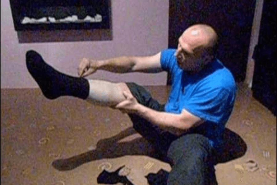 The image shows a man sitting on the floor attempting to put on multiple pairs of socks over one leg. He is in a casual setting, wearing a blue shirt and black socks, with additional socks scattered on the floor around him. The scene suggests he is engaged in a challenge or record attempt to wear the most socks at once, highlighting determination and humor.