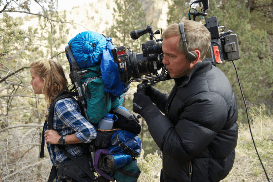 This image shows a woman wearing a large hiking backpack, dressed in a plaid shirt, walking outdoors in a forested area. Beside her, a man with headphones and gloves operates a professional camera, capturing her movement in what appears to be a behind-the-scenes moment from a film or documentary production.