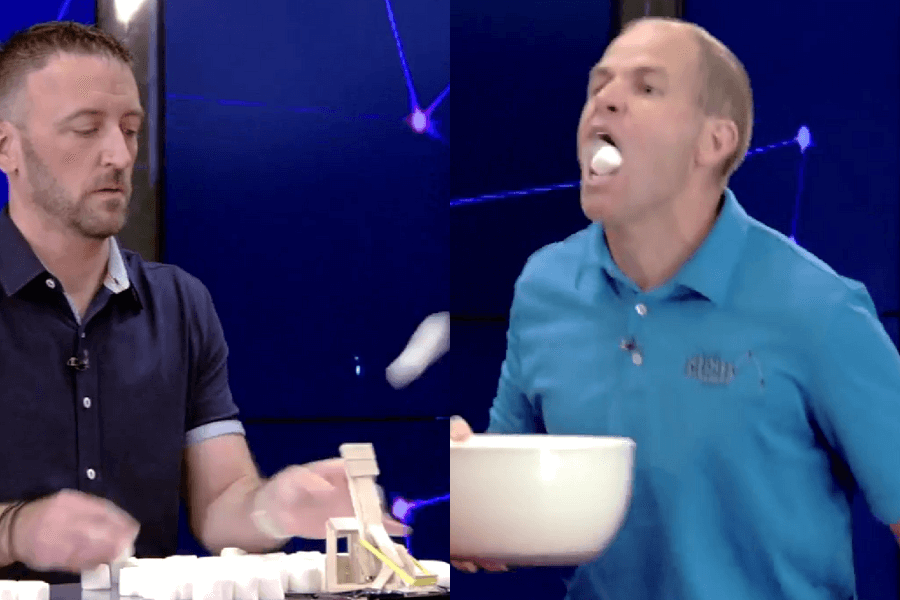 The image shows a split view of two men participating in a marshmallow-related challenge on a game or TV show. On the left, one man is setting up marshmallows with a small catapult-like device, appearing focused on launching them. On the right, the other man is catching marshmallows in his mouth, with an open bowl in his hands, showcasing excitement and effort. The colorful, futuristic backdrop adds an energetic atmosphere to the lighthearted and competitive activity.