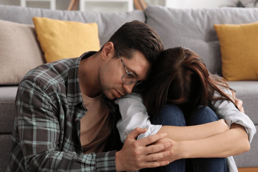 A man wearing glasses and a plaid shirt sits closely beside a woman who has her head down on her knees, offering comfort and support. They are seated on the floor in a cozy living room with a sofa and cushions in the background.