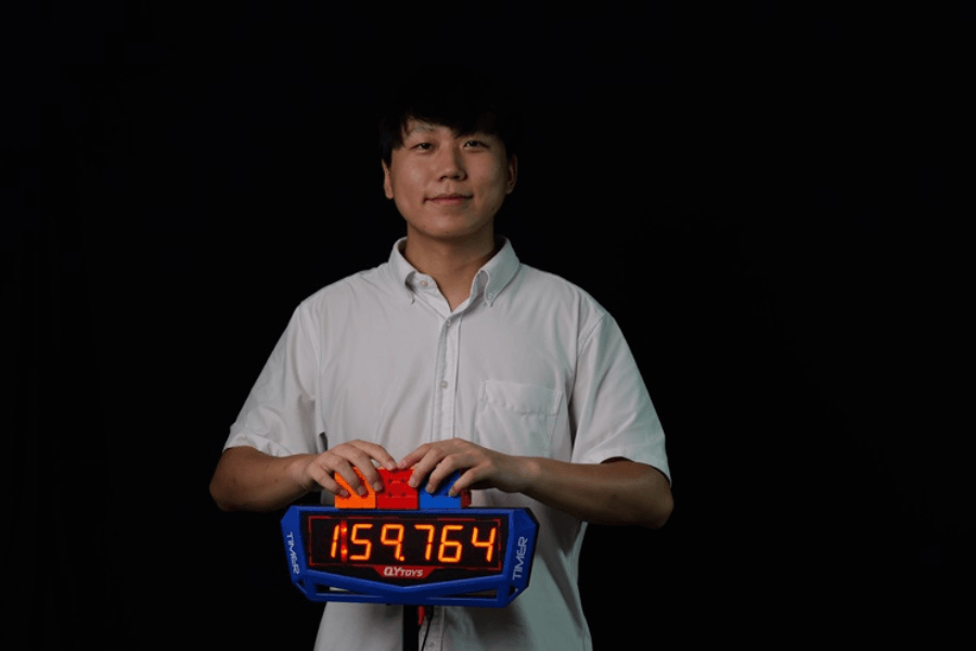 The image shows a young man proudly holding a timer device displaying the time "1:59.764," indicating a completed challenge or record attempt. He is dressed in a white shirt and stands against a dark background, emphasizing the timer and his achievement. The timer's design and his calm demeanor suggest this could be related to a speed-solving event, such as solving a Rubik's Cube or a similar timed task.