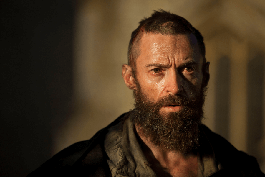 This image depicts a man with a rugged appearance, including a shaved head with scars, a thick beard, and intense eyes. He is wearing weathered clothing and stands in dramatic lighting, suggesting a gritty or emotional scene in a historical or dramatic context.