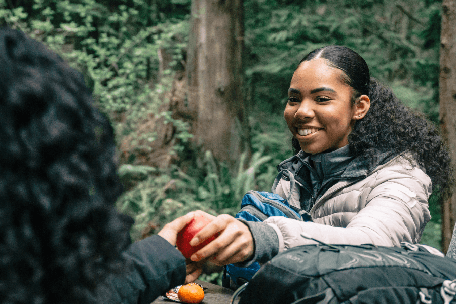 A woman wearing a puffy jacket smiles warmly as she hands a red apple to someone in a forest setting. Backpacks and oranges are visible, suggesting a relaxed outdoor hike or picnic.