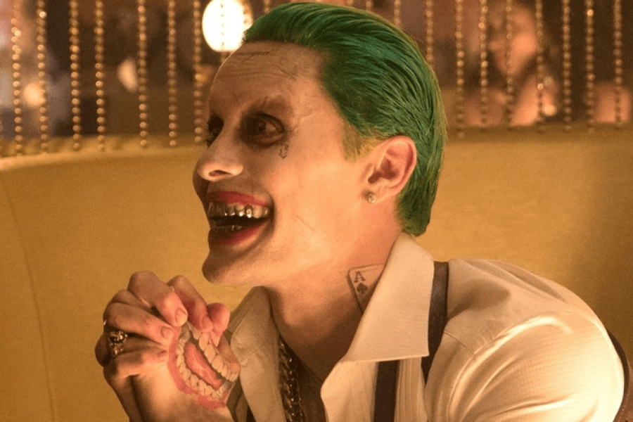 This image depicts a character with green hair, pale skin, red lips in a sinister smile, and prominent tattoos, wearing a white shirt and purple suspenders. The character's menacing grin reveals metallic teeth, adding to their theatrical, villainous appearance.