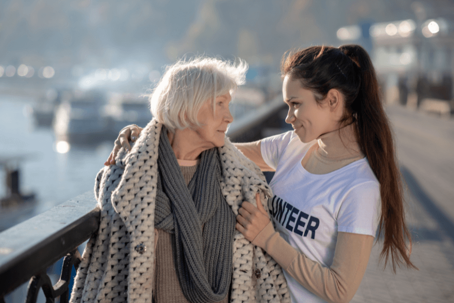 A young woman wearing a "Volunteer" shirt smiles warmly at an elderly woman wrapped in a thick knitted scarf and coat, as they stand together by a waterfront railing on a sunny day.