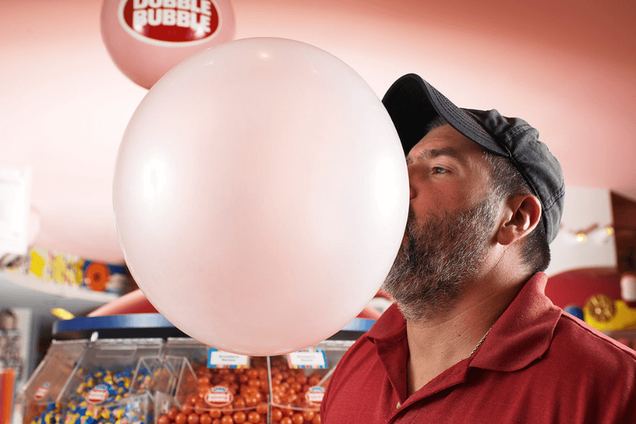 The image shows a man in a red shirt and black cap blowing an impressively large pink bubble gum bubble. The background features colorful candy bins and a "Dubble Bubble" logo, suggesting the setting is a candy store or a gum-related event. The bubble is nearly as large as the man's face, highlighting his bubble-blowing skill.