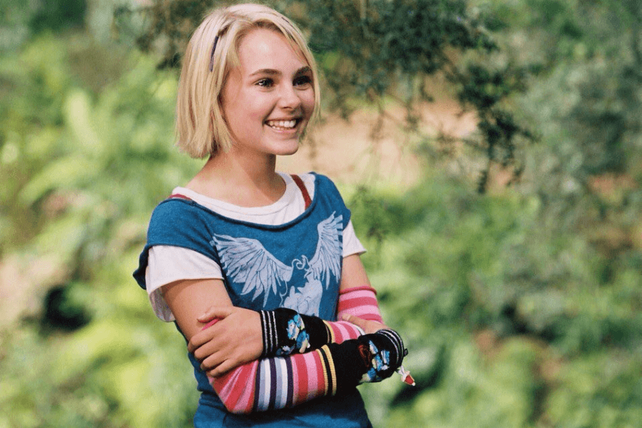 This image features a young girl with short blonde hair, smiling brightly while standing outdoors in a lush, green setting. She is wearing a blue shirt with a white angel-wing design, layered over a white top, and colorful striped arm warmers that add a playful touch. Her cheerful demeanor and vibrant outfit create an energetic and joyful atmosphere.