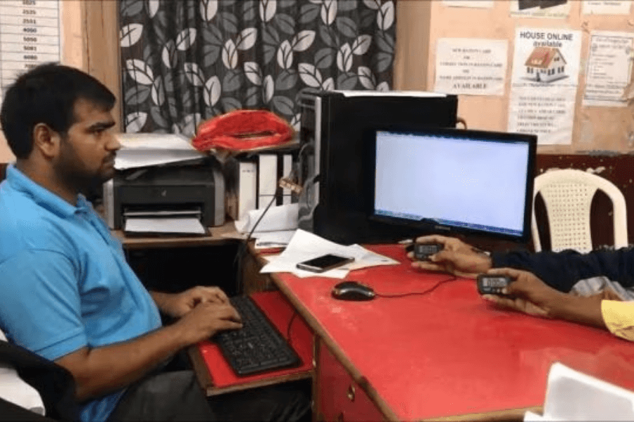 The image shows a man seated at a desk, typing on a keyboard, while another person holds two small digital counters or clickers. The setting appears to be an office with a computer monitor, documents, and a printer visible in the background. This scene likely depicts a task or challenge involving speed typing or data entry, possibly for a record attempt or efficiency demonstration. The focused atmosphere adds to the sense of purpose in the activity.