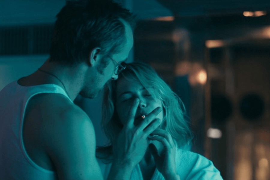 This image features an intimate moment between two characters under moody, blue-tinted lighting. A man in a tank top leans close to a woman who is smoking, with her eyes closed and an expression of contentment. The scene conveys a quiet, personal interaction in a dimly lit setting.