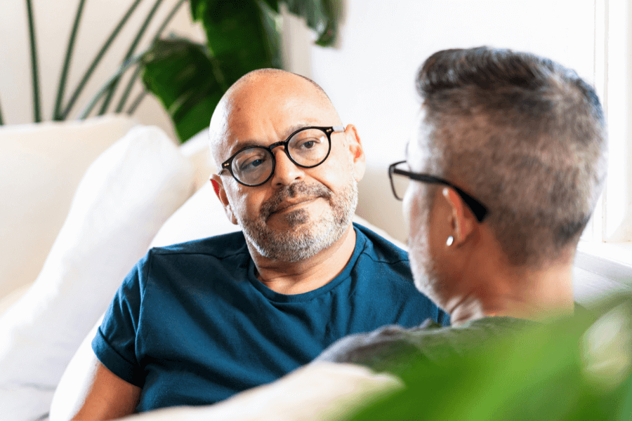 Two men sit on a couch in a relaxed indoor setting, engaged in a thoughtful conversation. One man, wearing glasses, listens attentively, while plants and soft lighting add to the cozy atmosphere.