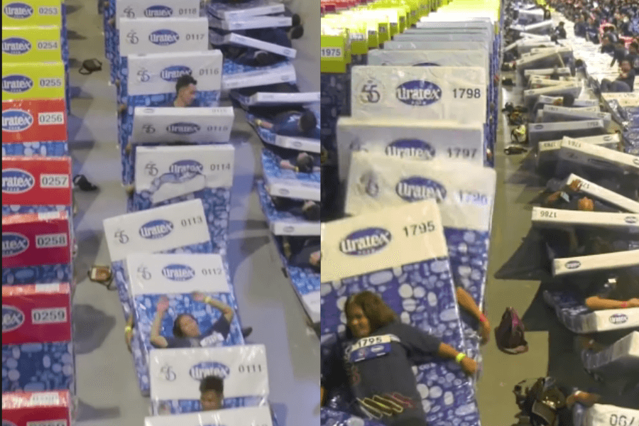 The image shows a large-scale domino effect involving people lying on mattresses, each labeled with numbers and branding from "Uratex." The participants are arranged in long rows, with the mattresses toppling over sequentially like dominos. The vibrant colors and organized setup suggest this is part of a Guinness World Records attempt for the largest human mattress domino. The event appears lively, with participants waving and engaging in the fun spectacle.